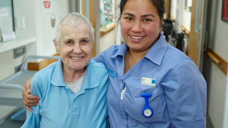 Aged care nurse with arm around elderly woman in a rest home corridor.