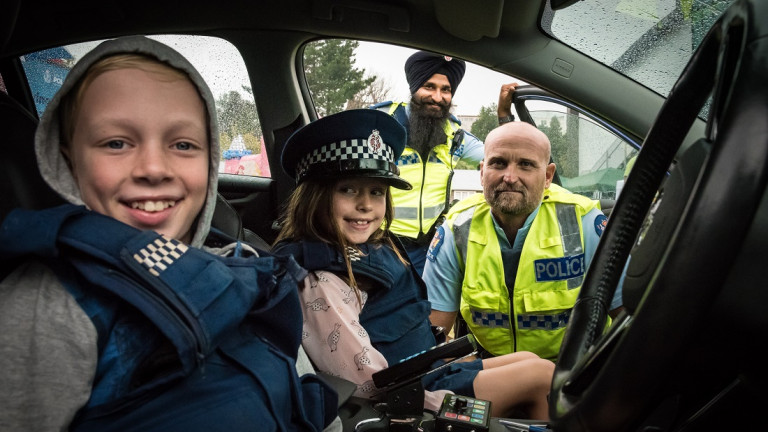 Children wearing police uniform hat and vest sitting in a car with two police men in background smiling