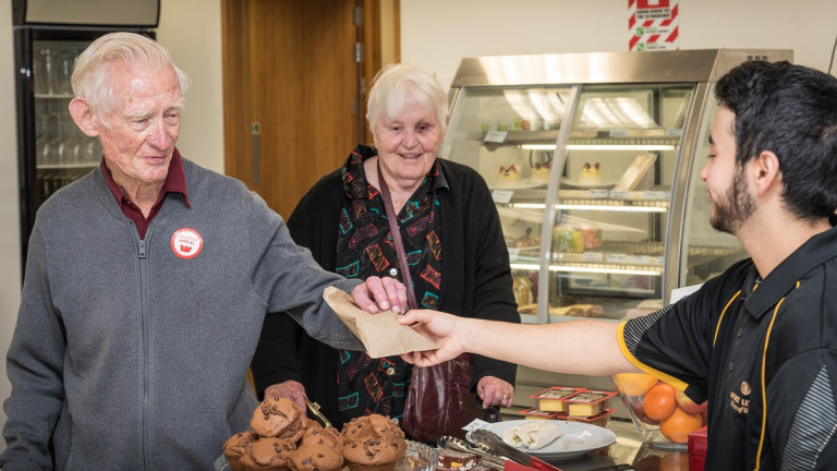 Elderly man and woman buying muffins from a rest home cafe worker.