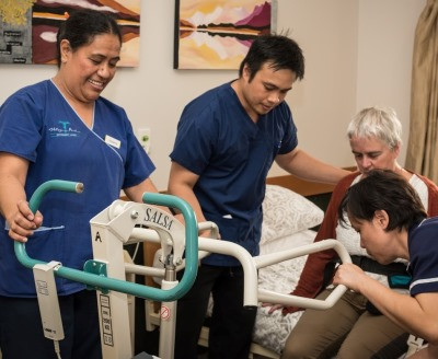 3 migrant aged care workers helping a woman into a hoist.