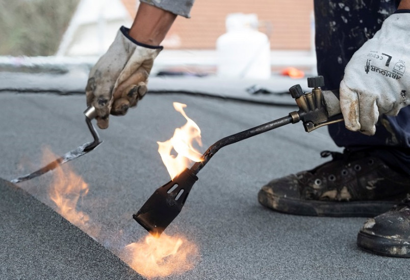 Construction worker wearing safety gloves and boots while using a blow torch to seal roofing