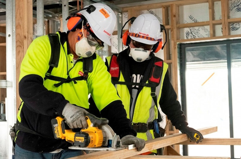 Two carpenters wearing helmets, ear muffs, face masks and gloves while cutting wood