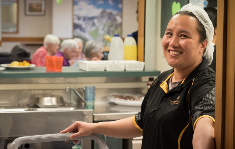 Migrant retirement village kitchen worker with a trolley wearing a hair net (elderly ladies eating in meal room in background)